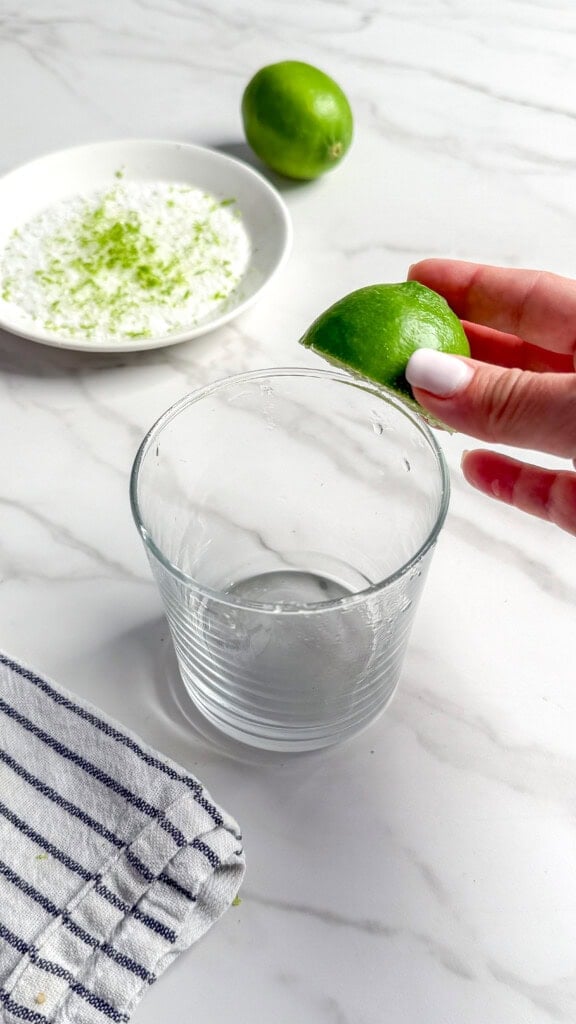 A hand with white nail polish squeezes a lime wedge over an empty glass on a white marble surface, capturing the fresh ingredients perfect for Classic Margaritas. In the background, whole limes, lime zest, and a striped cloth complete the scene.