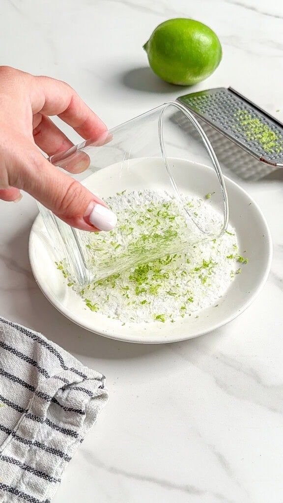 A hand presses a glass into a white plate with sugar and lime zest—perfect prep for Fresh Classic Margaritas. A whole lime, zester, and striped towel rest nearby on a white countertop.