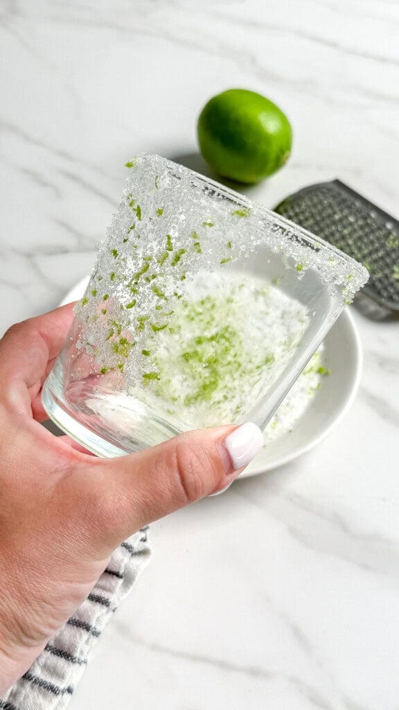 A hand holds a glass rimmed with sugar and lime zest above a bowl, ready for Fresh Classic Margaritas. In the background, there’s a whole lime and a grater on a marble countertop.