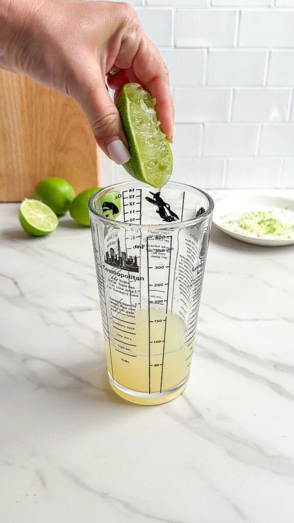 A hand squeezes a halved lime over a clear measuring glass, collecting juice for Fresh Classic Margaritas. In the background are more limes, a plate with lime zest, and a wooden cutting board on a white kitchen countertop.