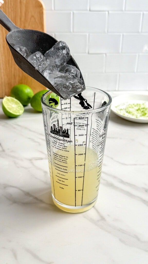 A metal scoop pours ice into a glass measuring cup filled with yellow liquid on a marble counter, ready for Fresh Classic Margaritas. In the background, limes—one sliced and one whole—rest on a wooden cutting board.