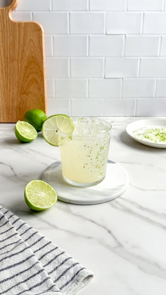 A glass of lime drink with ice and a lime wheel garnish sits on a round white coaster. Fresh limes, a plate with zest, and a striped towel surround the scene—perfect ingredients for crafting Fresh Classic Margaritas on the marble countertop.