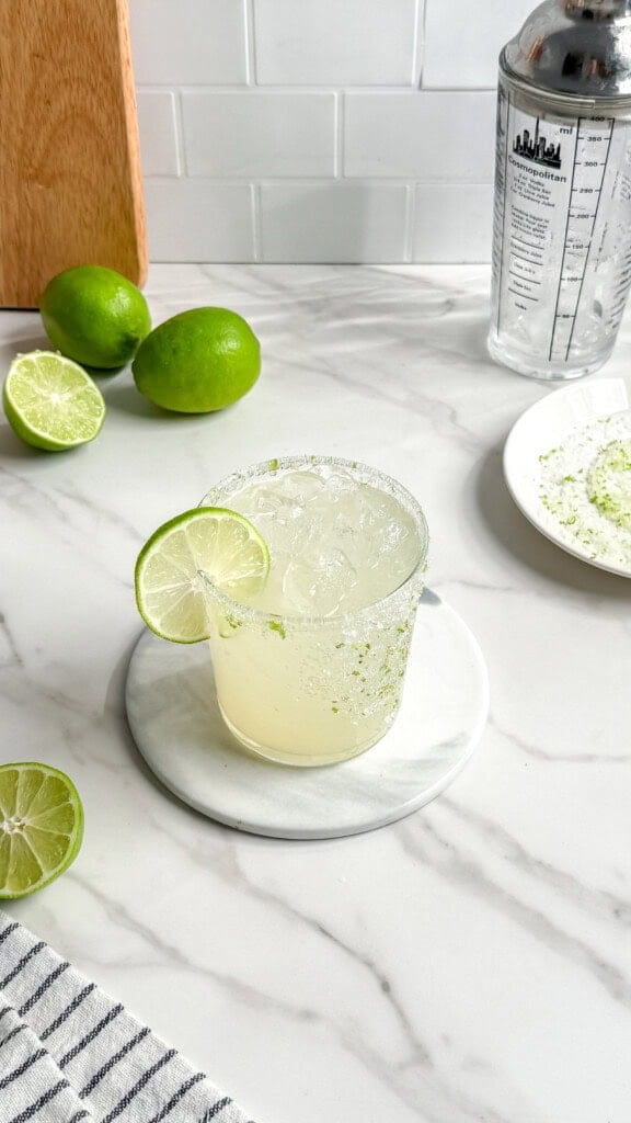 A glass of Fresh Classic Margaritas with ice and a lime wheel garnish sits on a marble coaster, surrounded by sliced and whole limes, a cocktail shaker, and a plate with salted lime zest on a white marble countertop.