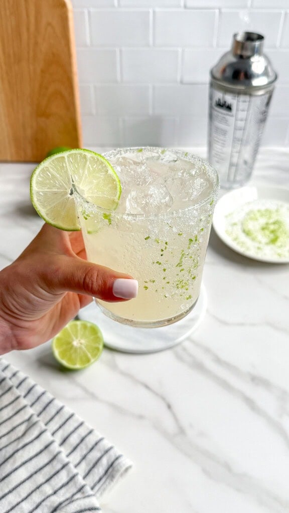 A hand with manicured nails holds a glass of Fresh Classic Margaritas, its salted rim and lime wheel garnish gleaming. A cocktail shaker, lime slices, and a striped towel rest on the white marble countertop in the background.