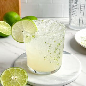 A refreshing Fresh Classic Margarita with a salted rim and a lime wheel garnish sits on a marble coaster. Halved limes, a plate with salt, and a cocktail shaker complete the scene on a white countertop.