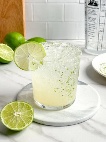 A refreshing Fresh Classic Margarita with a salted rim and a lime wheel garnish sits on a marble coaster. Halved limes, a plate with salt, and a cocktail shaker complete the scene on a white countertop.