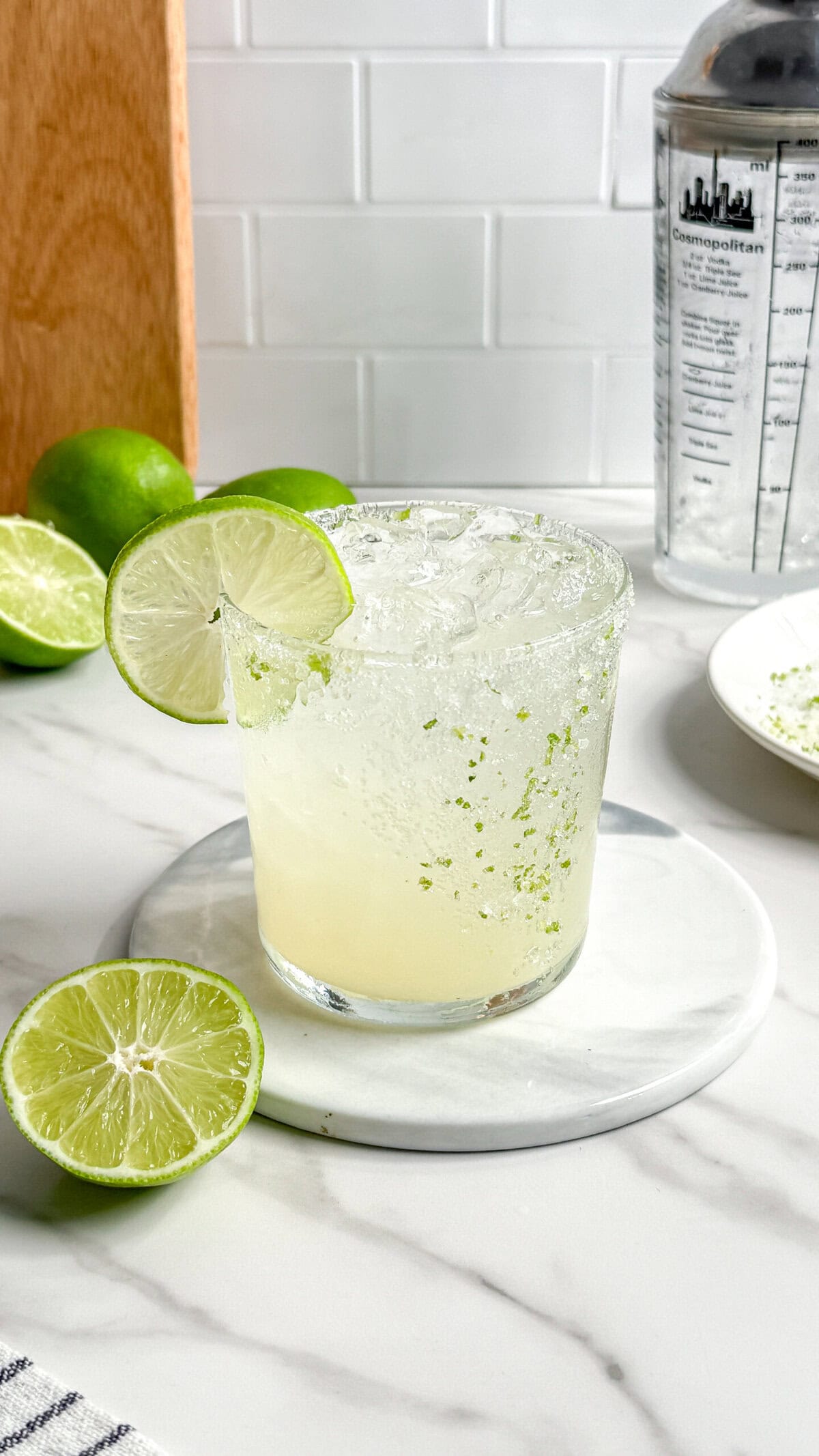A refreshing Fresh Classic Margarita with a salted rim and a lime wheel garnish sits on a marble coaster. Halved limes, a plate with salt, and a cocktail shaker complete the scene on a white countertop.