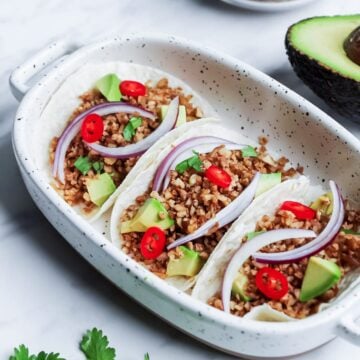 Three tacos in a white dish filled with vegan walnut taco meat, diced avocado, tomato, and sliced onions with a white marble background.