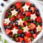 A red white and blue fruit salad featuring strawberries, blueberries, cherries, watermelon, and star-shaped melon pieces, garnished with fresh mint leaves and surrounded by extra fruit on a white surface.