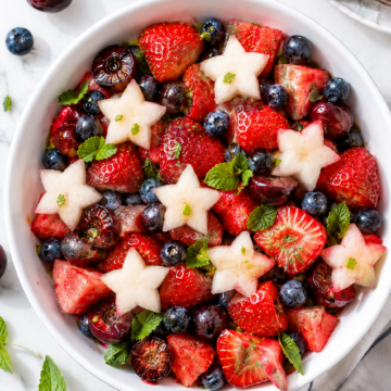 A red white and blue fruit salad featuring strawberries, blueberries, cherries, watermelon, and star-shaped melon pieces, garnished with fresh mint leaves and surrounded by extra fruit on a white surface.
