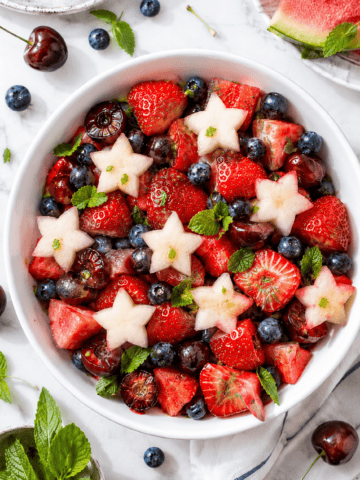 A red white and blue fruit salad featuring strawberries, blueberries, cherries, watermelon, and star-shaped melon pieces, garnished with fresh mint leaves and surrounded by extra fruit on a white surface.
