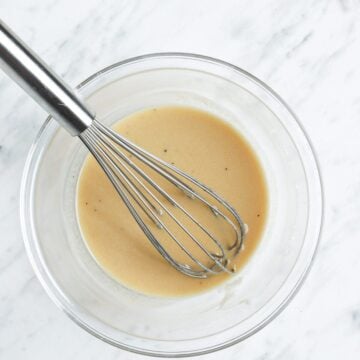 Mediterranean tahini salad dressing in a glass bowl with a large whisk with a white marble background.