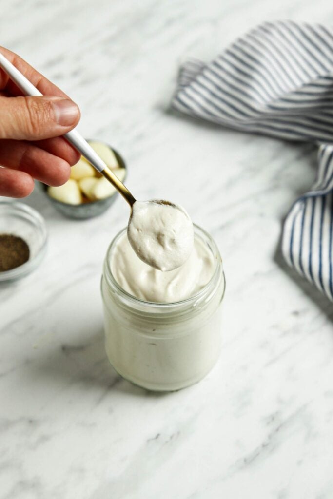 A glass jar of easy vegan pasta sauce with a silver spoon lifting sauce above it next to a striped towel with a white marble background.