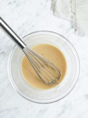 Mediterranean tahini salad dressing in a glass bowl with a large whisk with a white marble background.