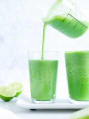A green detox smoothie being poured from a glass pitcher into a glass cup with a white background.