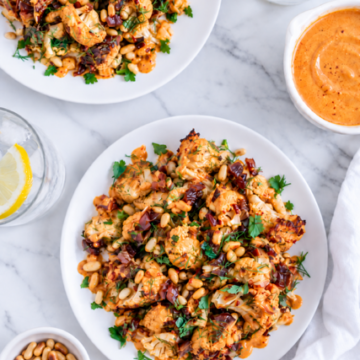 Two plates of Harissa Cauliflower salad with pine nuts, parsley, and spices sit on a marble surface. A bowl of pine nuts, a dish of orange sauce, and glasses of water with lemon are nearby.