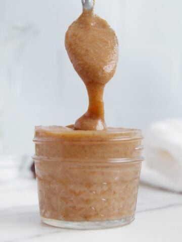 A spoonful of vegan caramel sauce dripping into a glass container of vegan caramel sauce.