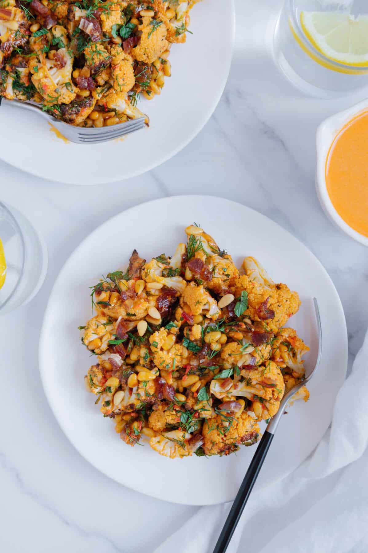 Roasted cauliflower tossed with dates, pine nuts and harissa tahini sauce on a white plate served with a black fork.