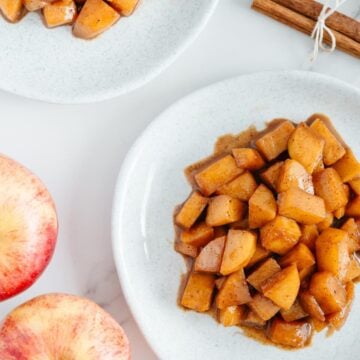 Apple compote on two white plates next to two whole red apples and cinnamon sticks with a white marble background.