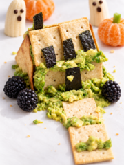 Spooky Halloween snack shaped like a haunted house, inspired by classic Halloween Haunted Houses, made of crackers, avocado, and seaweed, with blackberries, banana ghosts, and clementine pumpkins in the background.