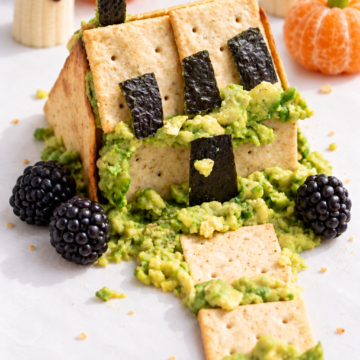 Spooky Halloween snack shaped like a haunted house, inspired by classic Halloween Haunted Houses, made of crackers, avocado, and seaweed, with blackberries, banana ghosts, and clementine pumpkins in the background.