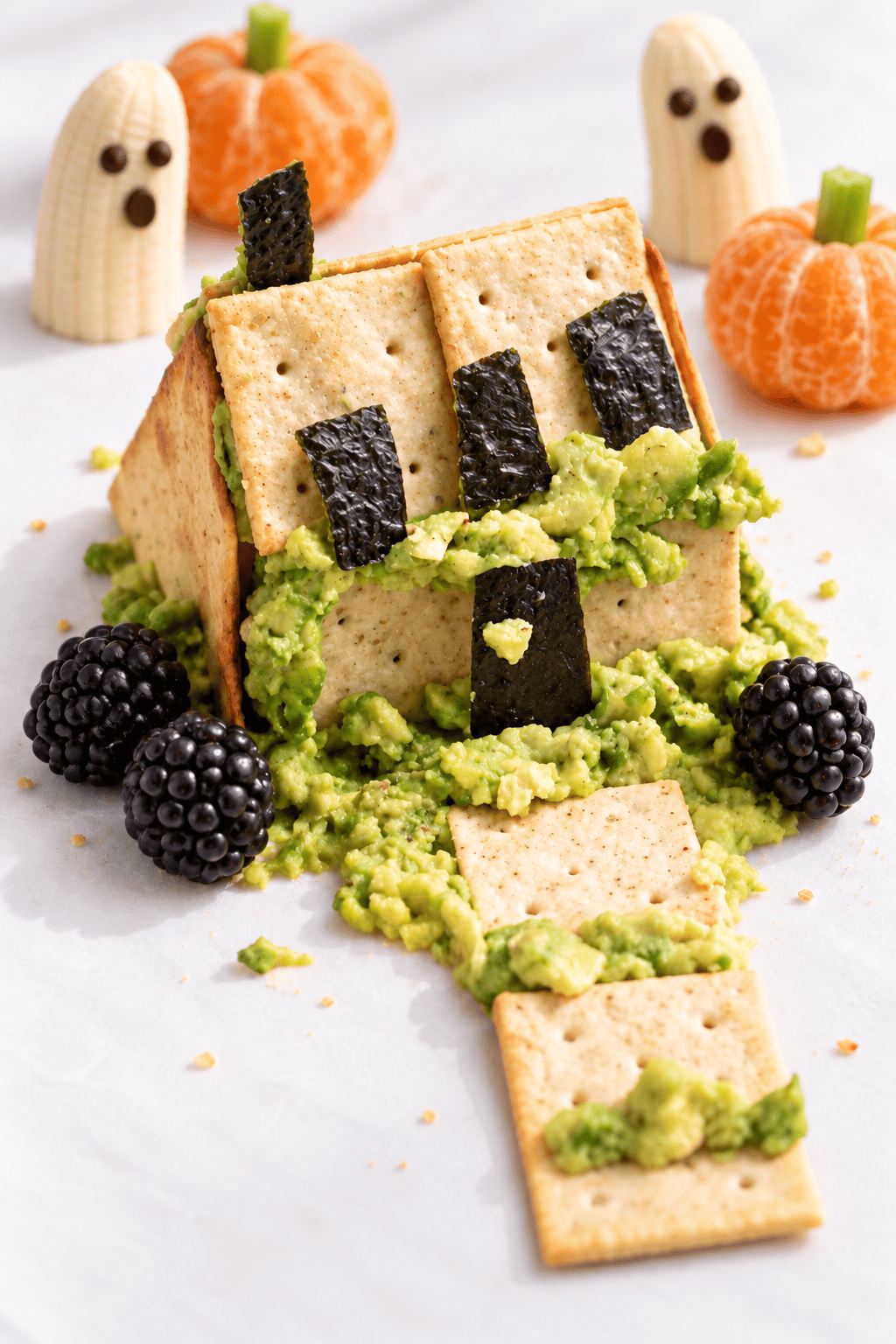 Spooky Halloween snack shaped like a haunted house, inspired by classic Halloween Haunted Houses, made of crackers, avocado, and seaweed, with blackberries, banana ghosts, and clementine pumpkins in the background.