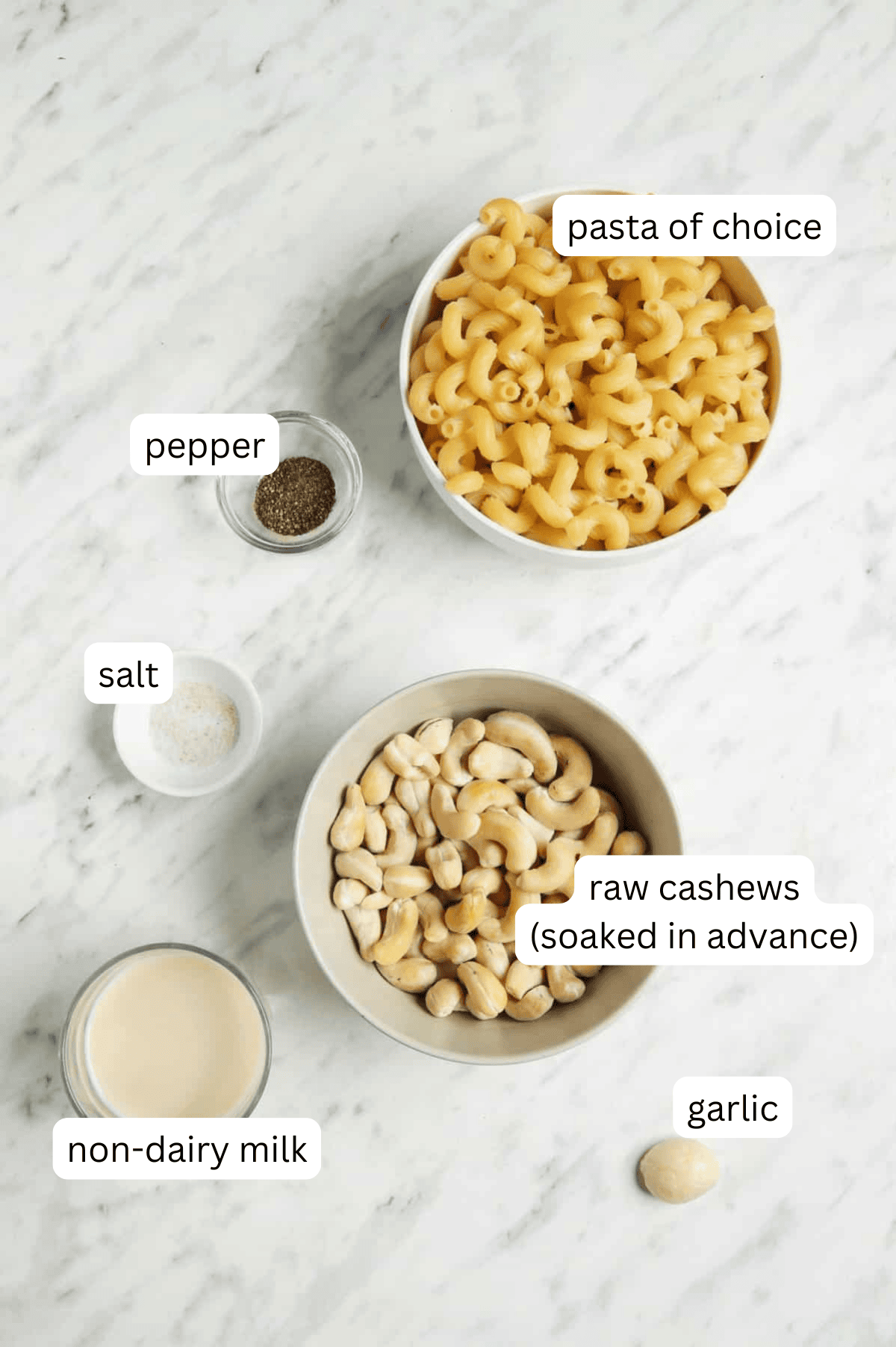 Ingredients of vegan cashew pasta sauce in separate bowls with a white marble background.
