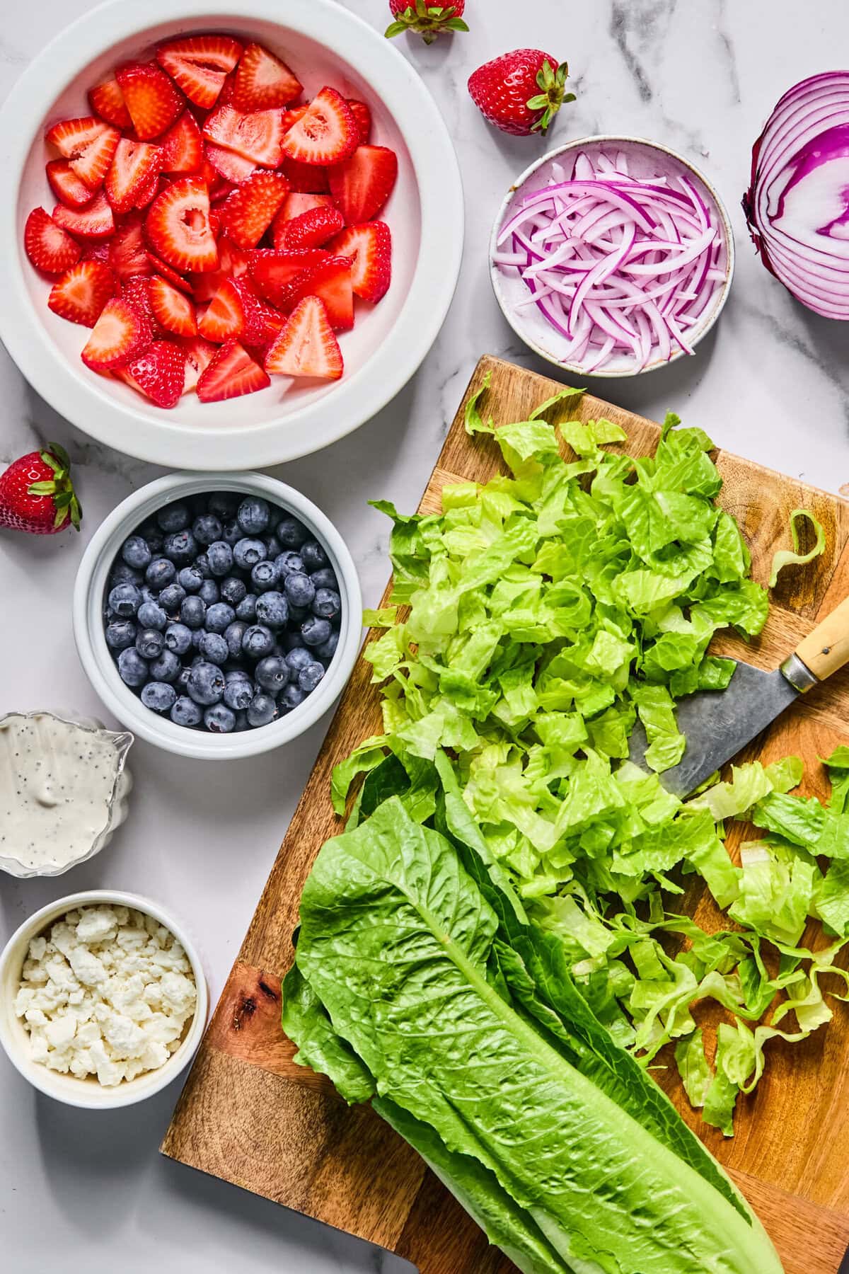 A cutting board with chopped romaine lettuce, a knife, and whole lettuce sits beside bowls of sliced strawberries, blueberries, red onions, crumbled cheese, and creamy dressing—perfect for making a Red White and Blueberry Salad on a white surface.