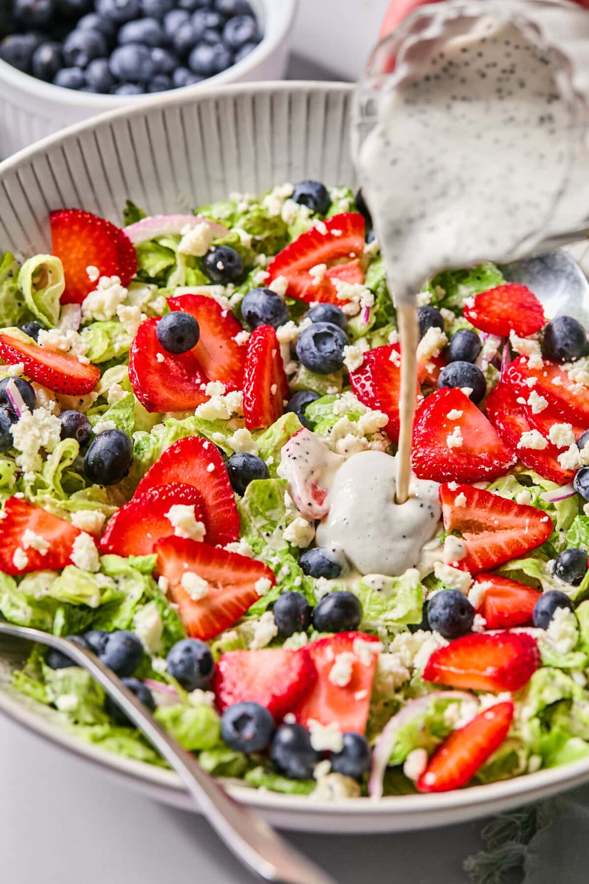 A fresh Red White and Blueberry Salad with lettuce, sliced strawberries, blueberries, red onion, and crumbled cheese is in a bowl. Creamy poppy seed dressing is being poured over the salad. A bowl of blueberries sits in the background.
