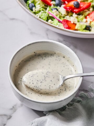 A bowl of creamy poppy seed dressing with a spoon rests on a light surface. In the background, a vibrant Red White and Blueberry Salad with lettuce, blueberries, strawberry slices, and feta cheese is partially visible.