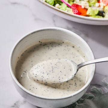 A spoonful of creamy, speckled dressing is held over a small white bowl, with a vibrant Red White and Blueberry Salad in the background on a marble surface.