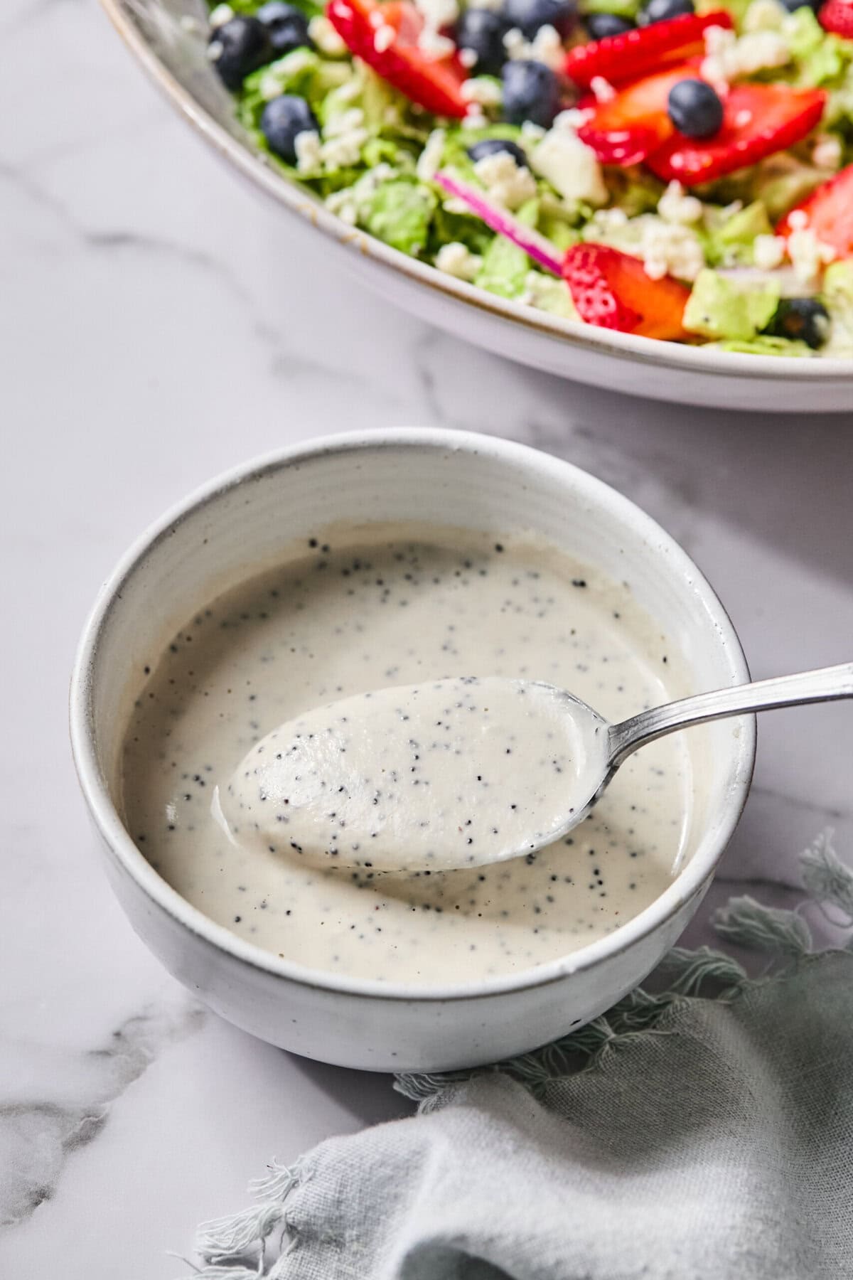 A spoonful of creamy, speckled dressing is held over a small white bowl, with a vibrant Red White and Blueberry Salad in the background on a marble surface.
