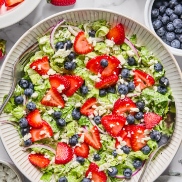 A fresh Red White and Blueberry Salad with chopped lettuce, sliced strawberries, blueberries, red onion, and crumbled cheese is served in a large bowl with serving spoons. Surrounding the bowl are extra berries and a bowl of dressing.