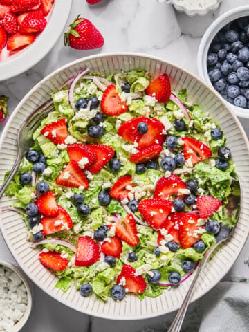 A fresh Red White and Blueberry Salad with chopped lettuce, sliced strawberries, blueberries, red onion, and crumbled cheese is served in a large bowl with serving spoons. Surrounding the bowl are extra berries and a bowl of dressing.