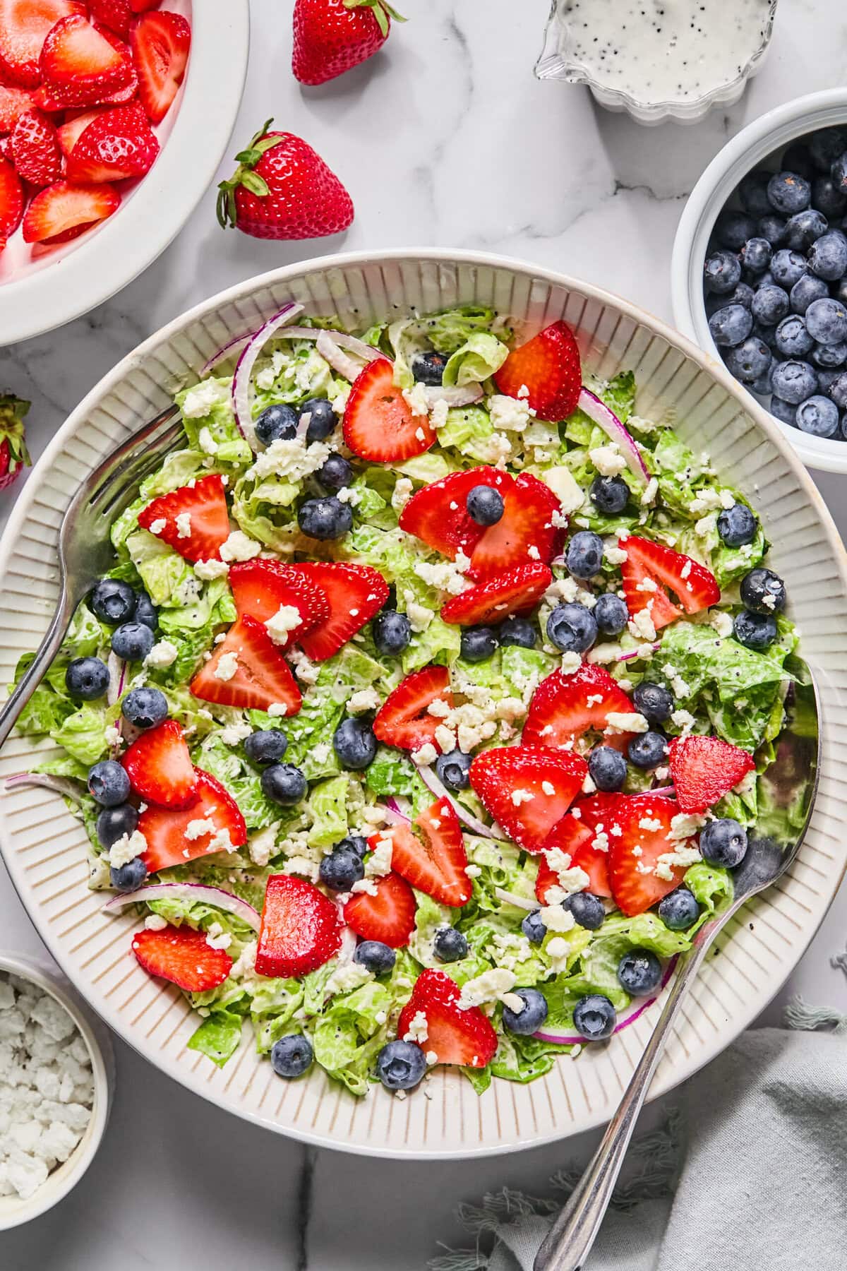 A fresh Red White and Blueberry Salad with chopped lettuce, sliced strawberries, blueberries, red onion, and crumbled cheese is served in a large bowl with serving spoons. Surrounding the bowl are extra berries and a bowl of dressing.