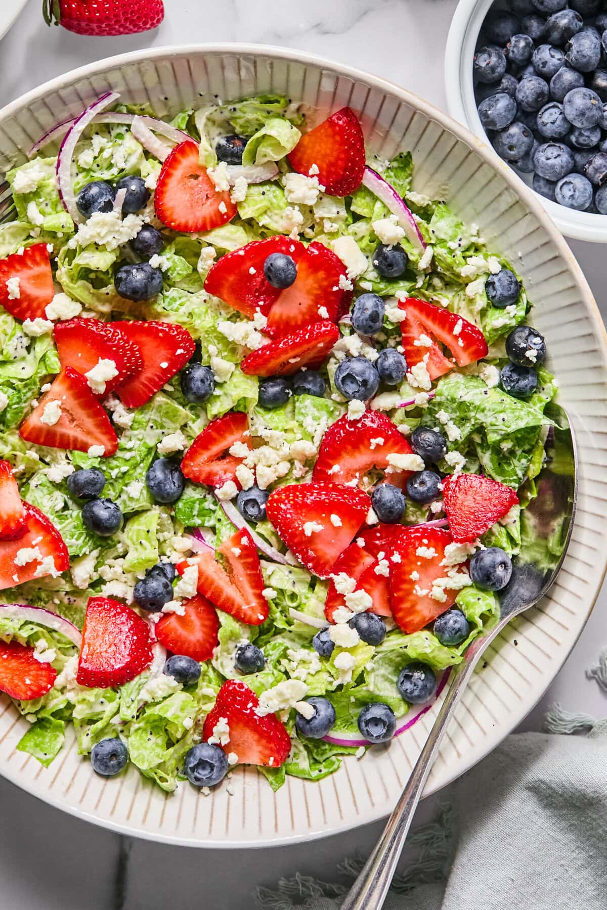 A plate of fresh Red White and Blueberry Salad with sliced strawberries, whole blueberries, lettuce, red onion, and crumbled cheese, with a spoon on the plate. A small bowl of blueberries and a strawberry are nearby.
