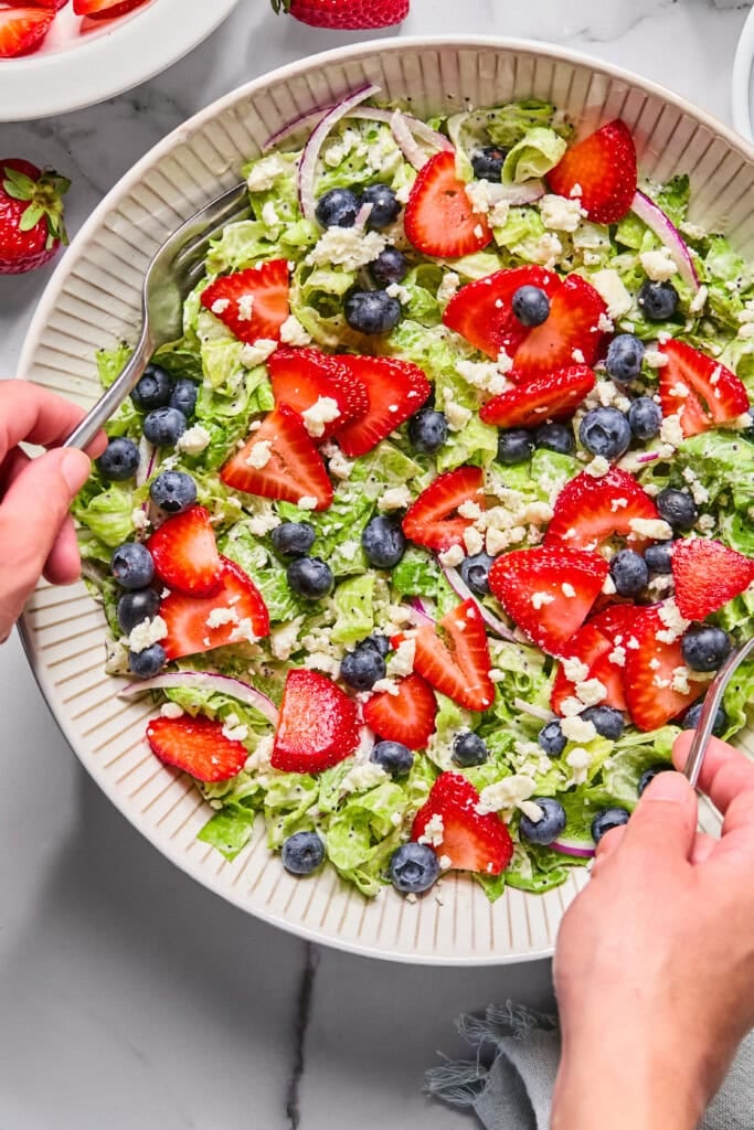 A fresh Red White and Blueberry Salad in a white bowl with sliced strawberries, blueberries, red onion, crumbled cheese, and lettuce. Two hands are tossing the salad with serving utensils.