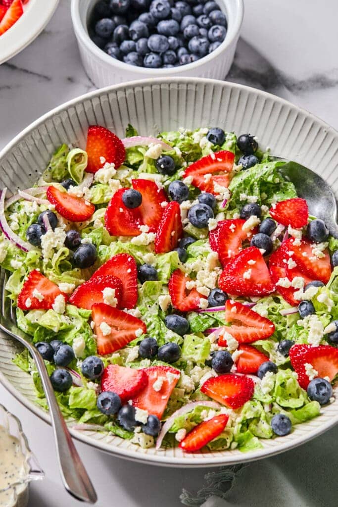 A bowl of Red White and Blueberry Salad topped with sliced strawberries, blueberries, crumbled cheese, and red onion slices sits ready with a spoon. A small dish of fresh blueberries is in the background.