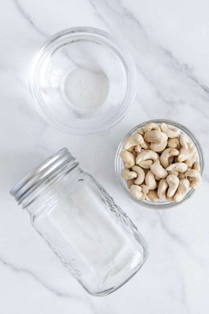 Overhead angle of raw cashews in a glass bowl next to an empty glass jar and a bowl of water.