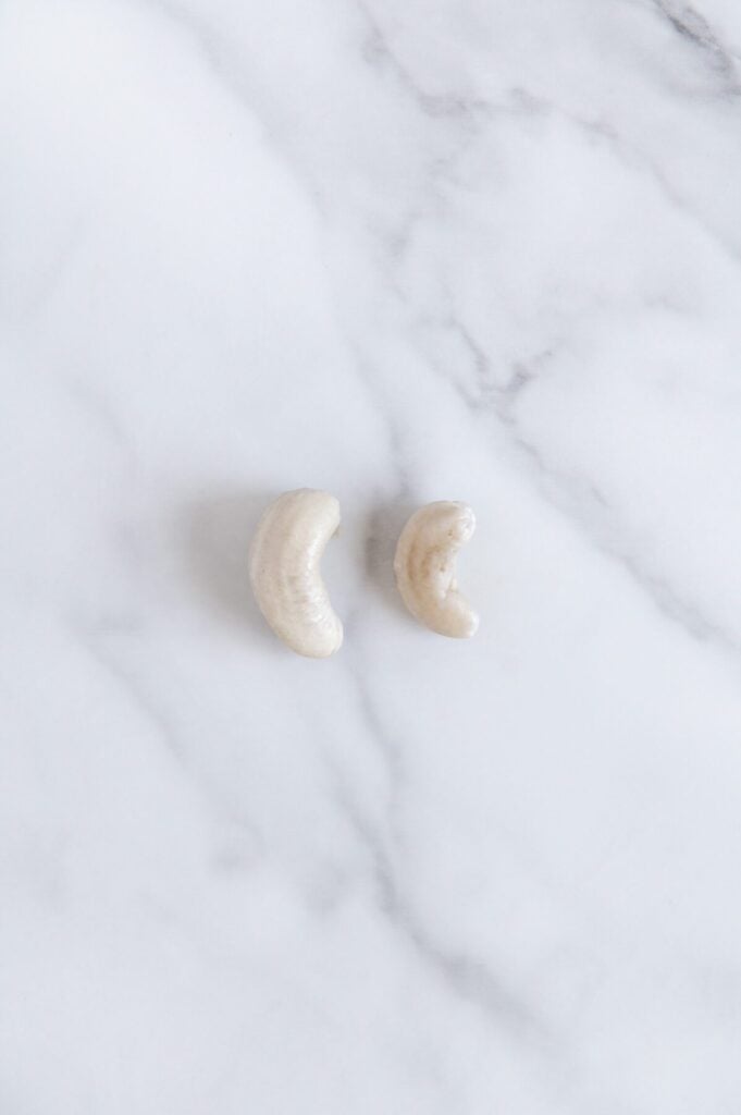 Overhead angle of a soaked cashew next to a raw cashew with a white marble background.