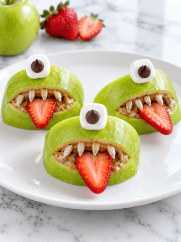 Three green apple slices made to look like monsters with strawberry tongues, sunflower seed teeth, marshmallow eyes, and chocolate chip pupils are arranged on a white plate with a strawberry in the background&mdash;perfect for pairing with other Spooky Spiders treats at your Halloween table.