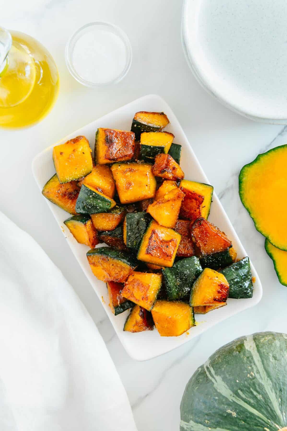 Maple sugar roasted Kabocha squash on a white platter next to olive oil and sliced Kabocha squash with a white background.
