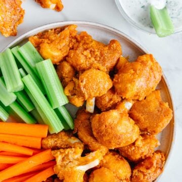 Buffalo cauliflower bites served on a plate with carrots and celery sticks next to a bowl of ranch dressing with a white marble background.
