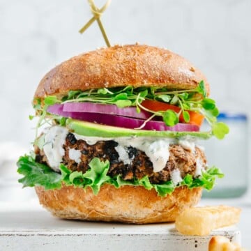 A grillable quinoa black bean veggie burger served on a bun with lettuce, tomato, avocado and vegan tzatziki with a white background.