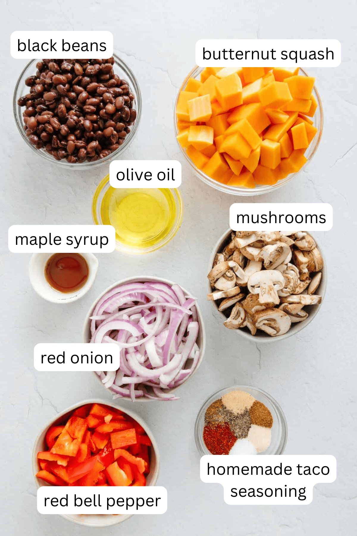 Top-down view of bowls with black beans, butternut squash, olive oil, mushrooms, red onion, red bell pepper, maple syrup, and taco seasoning arranged on a light surface-ideal ingredients for Sheet Pan Veggie Black Bean Tacos. Labels identify each item.