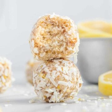 Two Lemon Coconut Energy Bites, one with a bite taken out, are stacked on each other, surrounded by shredded coconut and fresh lemon slices in the background.