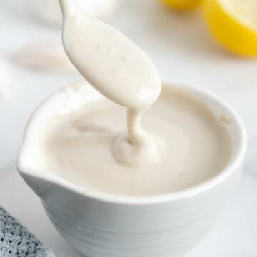 A spoonful of lemon garlic tahini dressing over a white bowl of tahini dressing next to lemons on a white surface.