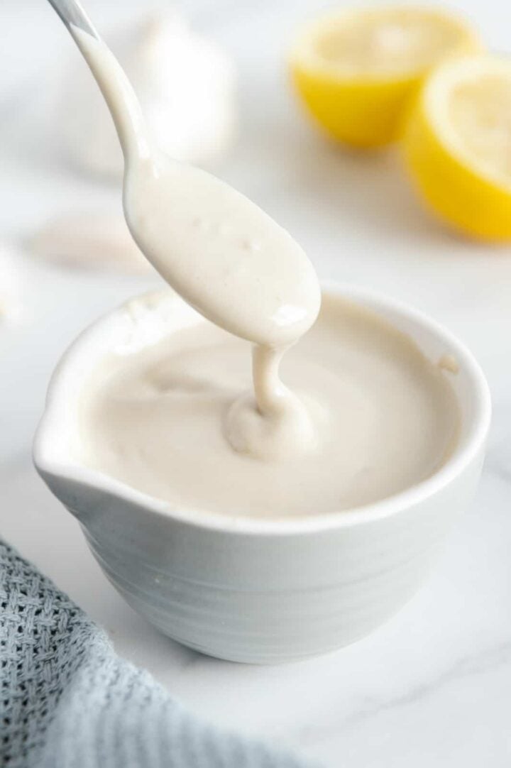 A spoonful of lemon garlic tahini dressing held over a small white bowl of dressing with a white background.