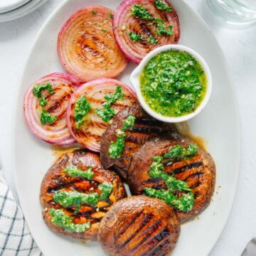 Grilled portobello mushrooms and sliced red onions topped with chermoula sauce on a white platter.
