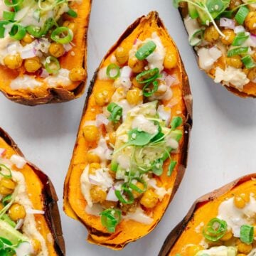 Five baked sweet potato halves are transformed in this Stuffed Sweet Potatoes Recipe, topped with roasted chickpeas, avocado chunks, microgreens, chopped red onion, green onions, and a light creamy drizzle on a white surface.